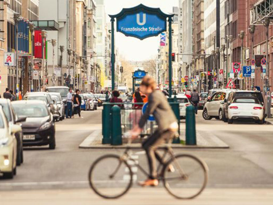 Fahrradfahrer auf belebter urbaner Kreuzung mit U-Bahn Station, Passanten und Autos